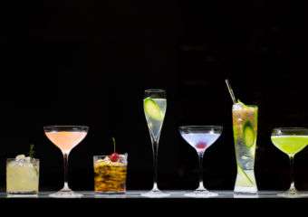 Five different cocktails displayed on a counter against black background.